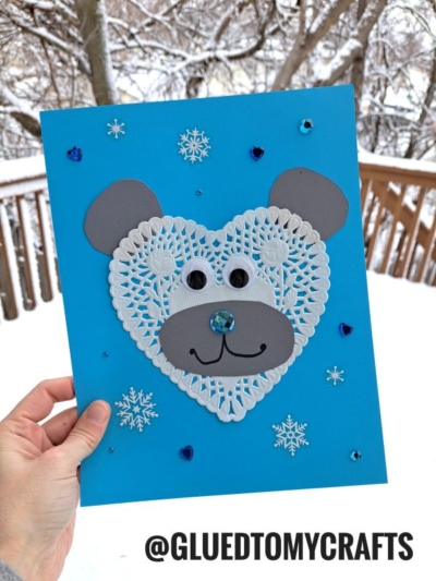 A hand holds a blue polar bear valentine card featuring a heart doily polar bear face with googly eyes and gray paper ears. Snowflake and heart stickers accent the card, with snowy trees visible in the background.