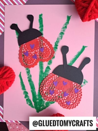 Two heart doily ladybug crafts made from red paper doilies, black accents, and purple hearts are displayed on a pink background with green paint resembling grass. The username @GLUEDTOMYCRAFTS appears at the bottom.