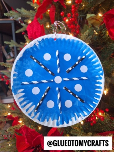 A paper plate snowflake ornament, adorned with white dot stickers and black-and-white striped straws, hangs on a Christmas tree decorated with red bows. The GluedToMyCrafts watermark is visible at the bottom.