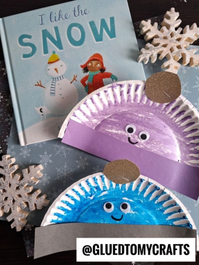 A children’s book titled I Like the Snow is surrounded by glittery snowflake decorations and two paper plate snowman crafts with painted faces, googly eyes, and a cute paper plate winter hat. The handle @GLUEDTOMYCRAFTS is at the bottom.