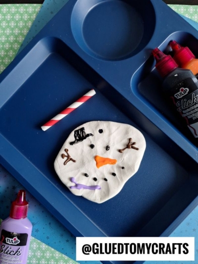 A blue tray holds a flattened white dough circle decorated as a snowman’s face with colored icing, resembling a melting snowman clay project, with a paper straw for the nose and bottles of black, orange, and purple puffy paint nearby. @GLUEDTOMYCRAFTS is at the bottom.