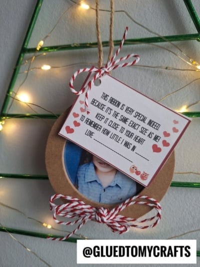A round height keepsake ornament with a child’s photo hangs from a green wire tree, adorned with a red and white string bow and a note reading, “This ribbon is very special indeed...” with heart and owl illustrations. Fairy lights glow in the background.