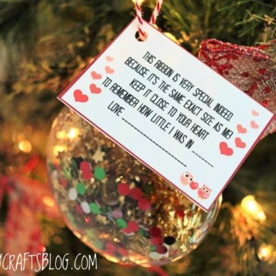 A handmade height keepsake ornament hangs on a Christmas tree. The ornament features a decorated card with hearts and bears, containing a sentimental message about measuring a child’s growth using ribbon.