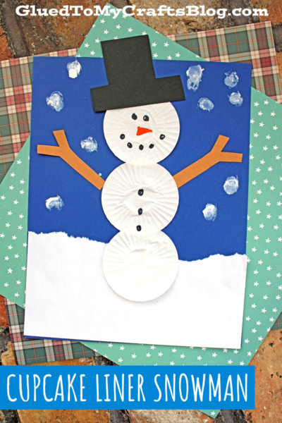 A snowman made from white cupcake liners, orange paper arms, and a black hat is glued on blue paper dotted with snowy white paint. Patterned scrapbook paper forms the background. Text reads, CUPCAKE LINER SNOWMAN—a fun cupcake liner snowmen craft!.