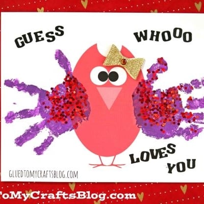 A red and pink handprint owl with googly eyes and a gold bow is decorated with two purple handprint wings on a white card reading GUESS WHO LOVES YOU. The red background features small gold hearts, making this a sweet handprint owl craft for kids.