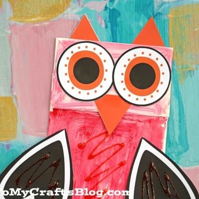 A handmade paper bag valentine owl puppet with heart-shaped wings and large round eyes, crafted from painted paper and craft materials, is displayed on a colorful background.