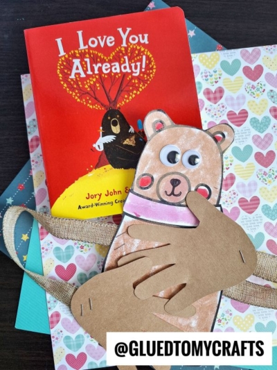 A red children’s book titled I Love You Already! featuring a bear on the cover rests on heart-patterned paper next to a handmade bear hug craft made from colored paper, with @GLUEDTOMYCRAFTS displayed at the bottom.