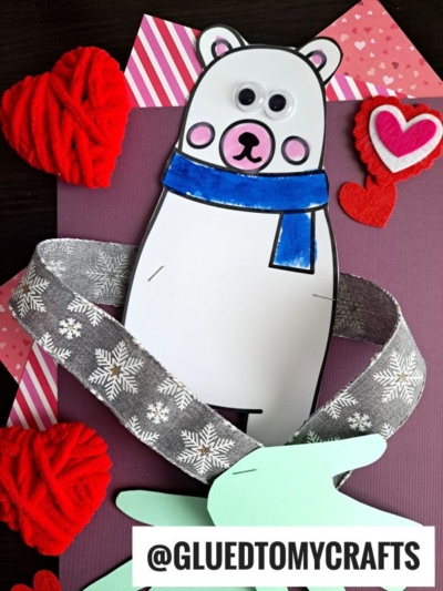 A handmade polar bear craft with googly eyes, a blue scarf, and red felt hearts surrounding it—perfect for sending a cozy bear hug. Pink striped paper and a gray snowflake ribbon decorate the background. Handle @GLUEDTOMYCRAFTS is visible at the bottom.
