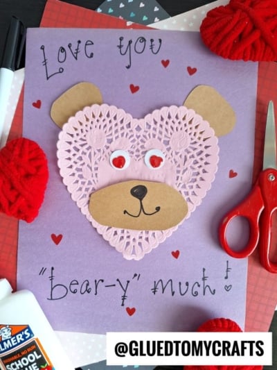 A handmade valentine bear card features a bear’s face crafted from a pink heart-shaped paper doily with red googly eyes, a brown paper snout and ears, and “Love you ‘bear-y’ much!” written on vibrant purple paper.