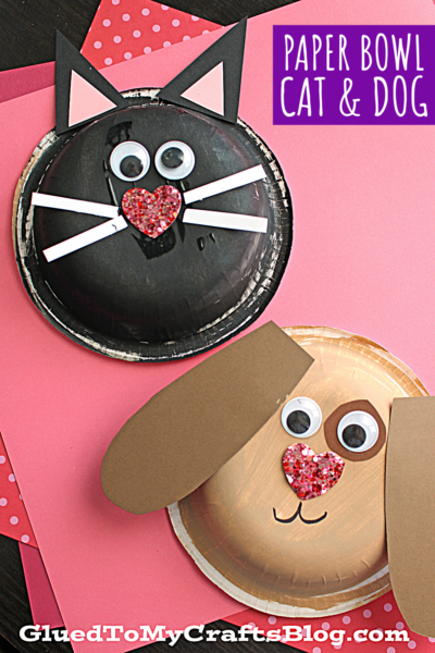 Two paper bowl crafts: a black cat with pointy ears, googly eyes, pink glitter heart nose, and whiskers, and a cute paper bowl puppy with floppy ears, googly eyes, and a pink glitter heart nose, on a pink background.