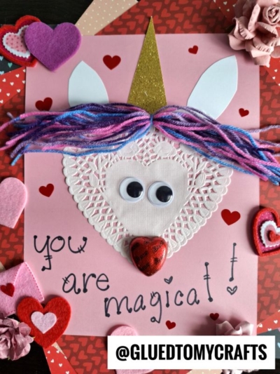 A handmade paper doily unicorn card with a gold horn, googly eyes, pink and purple yarn hair, felt hearts, and a heart-shaped nose. "You are magical!" is written on pink paper, surrounded by heart decorations.
