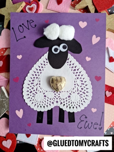 A handmade lamb valentine card features a sheep made from paper shapes, googly eyes, cotton balls, and a gold heart chocolate on a purple background with hearts. Text says “Love Ewe!” and “@GLUEDTOMYCRAFTS.”.