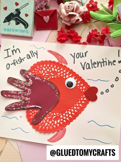 A handmade Valentine’s card features a heart doily fish with a paper handprint fin, a googly eye, and heart details. Text says, “I’m o-fish-ally your Valentine!” Surrounded by flowers and craft supplies.