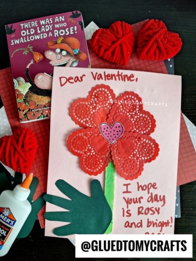 A handmade Valentine’s paper doily flower card with a green handprint stem and heart center sits next to red yarn roses, a glue bottle, scissors, and the book “There Was an Old Lady Who Swallowed a Rose!”.