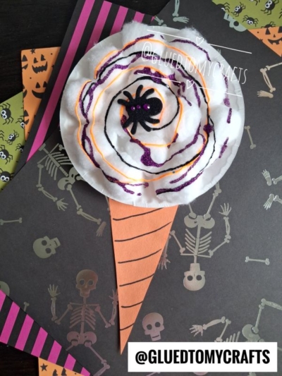 A Halloween craft shaped like an ice cream cone with a wavy, purple, orange, and black spider web scoop—just like a spooky cotton ball cotton candy treat. The background features skeleton and Halloween-themed paper. @GLUEDTOMYCRAFTS watermark is present.