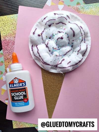 A paper cotton candy cone craft made with pink and brown paper, cotton balls, glitter, and purple glue sits next to a bottle of Elmer’s glue on a background of colorful papers. Text at the bottom: @GLUEDTOMYCRAFTS.