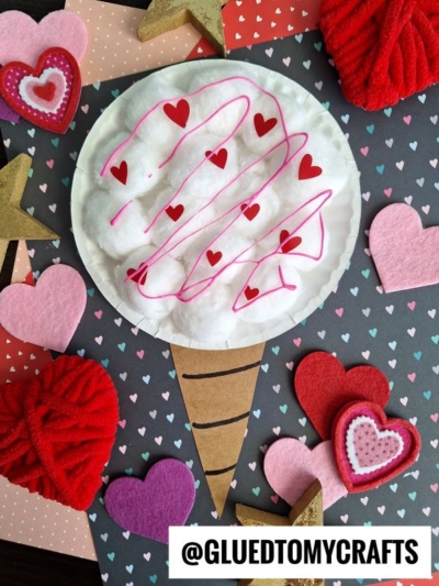 A paper cotton candy cone craft made with cotton balls, red heart stickers, and pink lines sits on a heart-patterned background, surrounded by felt and yarn hearts and gold stars. @GLUEDTOMYCRAFTS is printed at the bottom.