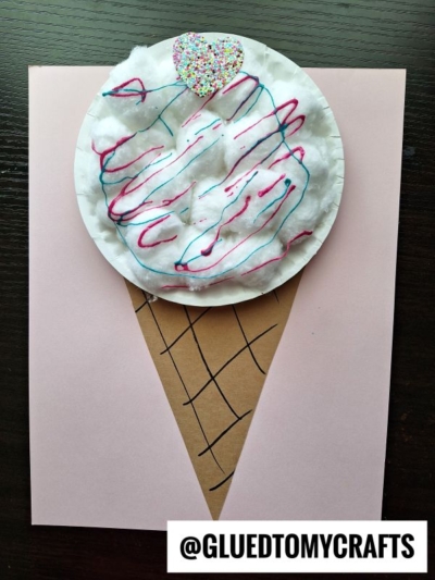 A paper craft ice cream cone made with a tan triangle for the cone, a white round paper plate resembling paper plate cotton candy topped with cotton balls and colored yarn for the ice cream, plus a heart-shaped sprinkle on top. @GLUEDTOMYCRAFTS is labeled below.
