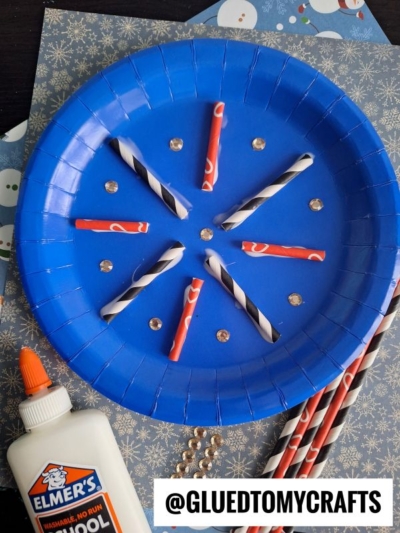 A blue paper plate snowflake is crafted with cut and glued striped paper straws, arranged in a snowy pattern and surrounded by small gems. Elmer’s glue and extra straws rest nearby on a snowflake-patterned background.