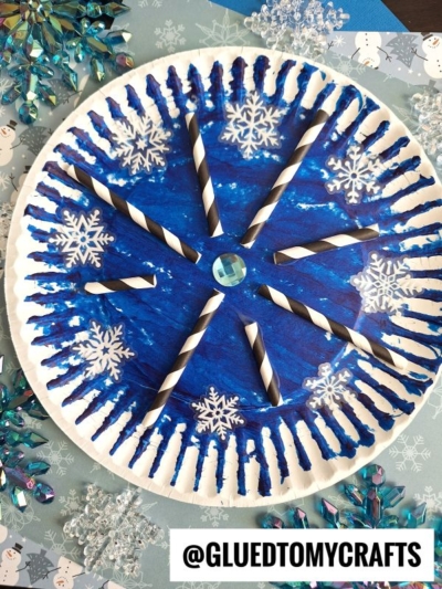 A blue and white paper plate snowflake craft, featuring black and white striped straws, snowflake cutouts, a central gem, and blue painted patterns. It’s beautifully displayed on a snowflake-themed background.