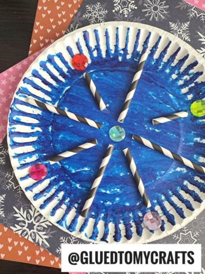 A paper plate snowflake painted blue with white streaks, decorated with black-and-white striped straws as spokes and colorful gem stickers at the ends, sits on patterned paper backgrounds. @GLUEDTOMYCRAFTS is written at the bottom.