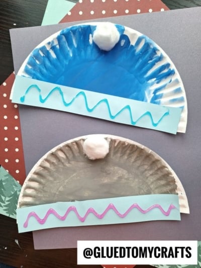 Two paper plate winter hat crafts painted blue and gray to look like cozy seasonal headwear, each decorated with a paper brim, a pompom on top, and a wavy line of paint across the brim, set on colored paper.