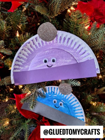 Two paper plate winter hat crafts resembling sparkly New Year’s Eve hats, decorated with googly eyes, colored paint, and construction paper, hang on a Christmas tree. The image includes the handle @GLUEDTOMYCRAFTS.