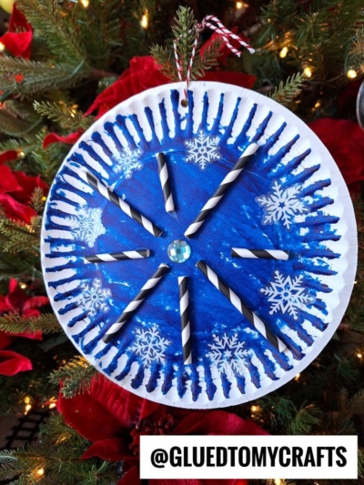 A blue paper plate snowflake ornament, decorated with black-and-white striped paper straws and a gem center, hangs on a Christmas tree surrounded by red poinsettias. @GLUEDTOMYCRAFTS is written at the bottom.