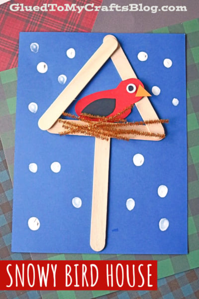 A blue paper showcases a mixed media snowy birdhouse made of craft sticks, a red bird cutout, and a brown pipe cleaner nest. White dots represent snow, and “Snowy Bird House” is written at the bottom.