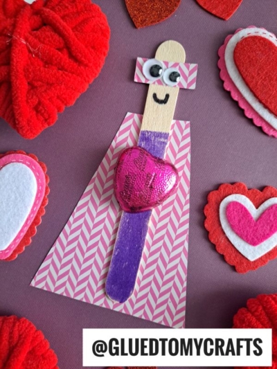 A popsicle stick valentine superhero craft with googly eyes, a marker-drawn smile, and a shiny heart on its chest, wearing a pink chevron cape and surrounded by red and pink felt hearts. Text reads @GLUEDTOMYCRAFTS.