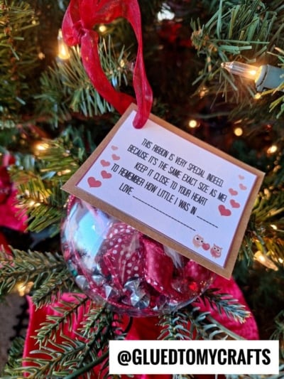 A Christmas height keepsake ornament with a red ribbon hangs on a tree. Attached is a note with hearts and owls: “This ribbon is very special indeed... to remember how little I was in love.” @GLUEDTOMYCRAFTS at the bottom.
