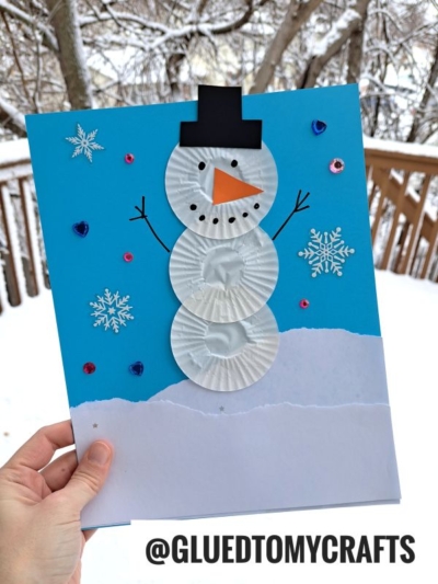 A hand holds a cupcake liner snowmen craft made from three white cupcake liners on blue paper, decorated with a black hat, orange nose, snowflake stickers, and sequins. Snowy trees and a wooden deck appear in the background.