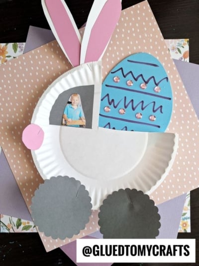 A handmade Easter craft featuring a bunny made from a paper plate, pink and white paper ears, a blue foam egg craft, and a photo of a child. The bunny’s wheels are gray circles, with patterned paper in the background.