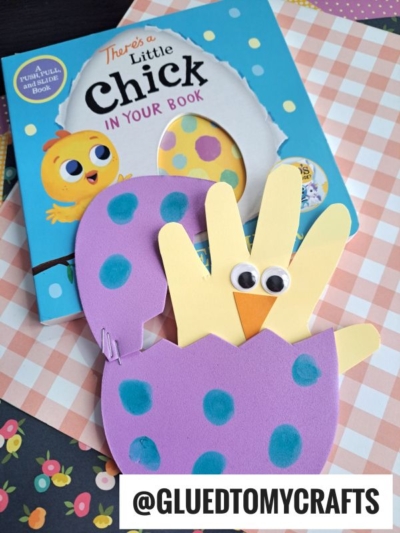 A children’s book titled Theres a Little Chick in Your Book sits behind a handmade chick craft made of yellow handprints, googly eyes, an orange beak, and a purple foam egg crafts shell with blue dots. The handle @GLUEDTOMYCRAFTS is visible.