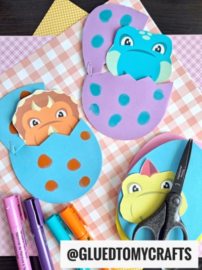 Colorful paper dinosaurs peek out of spotted foam egg crafts on patterned paper. Scissors and markers lie nearby, ready for creativity. A tag reads “@GLUEDTOMYCRAFTS.”.