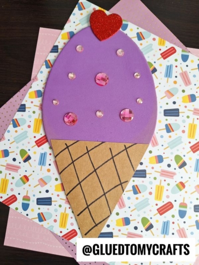A craft shaped like an ice cream cone with a brown paper cone, purple scoop, red heart at the top, and pink gems as decorations—perfect inspiration for foam egg crafts—placed on colorful patterned paper. @GLUEDTOMYCRAFTS is written at the bottom.