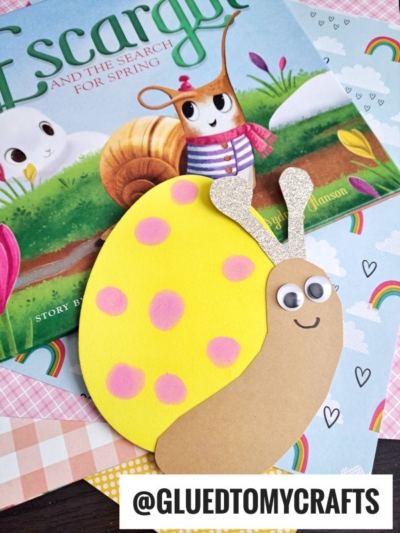 A handmade snail craft, made with foam egg crafts and featuring a yellow shell with pink dots, sits on patterned paper. Behind it is the book Escargot and the Search for Spring with illustrated snails. The tag @GLUEDTOMYCRAFTS is on the image.
