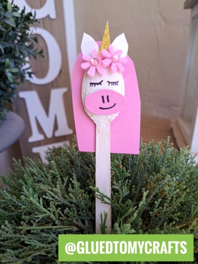 A wooden spoon unicorn, decorated with a pink mane, gold horn, flower crown, closed eyes, and smiling mouth, stands upright in a green plant. Text at bottom: @GLUEDTOMYCRAFTS.