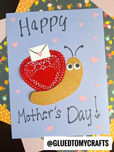 Handmade Mother’s Day card featuring a snail crafted from brown paper, a red paper doily heart shell, googly eyes, and a tiny envelope. “Happy Mother’s Day!” appears on blue paper with pink hearts—a sweet Paper Doily Crafts project.