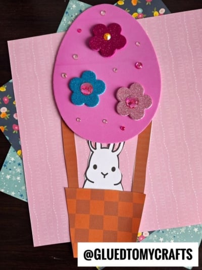 A handmade craft featuring a bunny peeking out from an orange basket below a large pink foam egg decorated with felt flowers and sequins, displayed on patterned paper backgrounds. @GLUEDTOMYCRAFTS is written at the bottom.