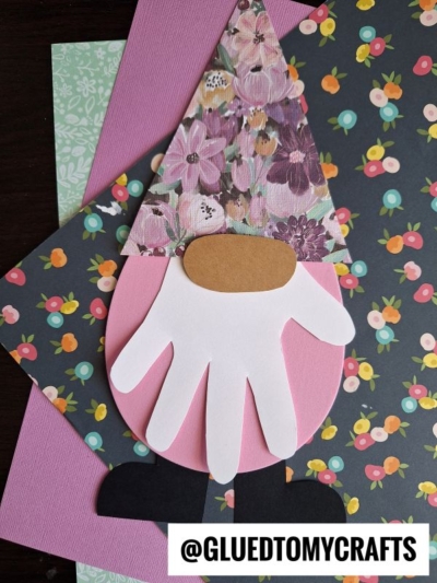 A handprint Easter gnome craft made with paper cutouts: a pink oval body, white handprint beard, floral hat, brown nose, and black boots, placed on colorful floral patterned paper. @GLUEDTOMYCRAFTS is displayed at the bottom.
