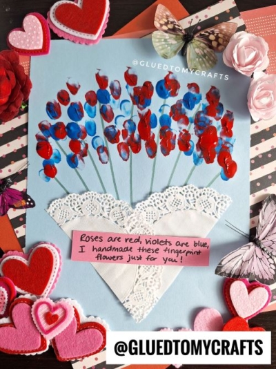A handmade craft featuring a light blue paper with a heart-shaped white doily as a vase, holding red and blue fingerprint flowers on green stems. The message reads, "Roses are red, violets are blue, I handmade these fingerprint flowers just for you!" The craft is surrounded by felt hearts, artificial flowers, and decorative butterflies, set against a patterned background. The watermark "@GLUEDTOMYCRAFTS" is visible.