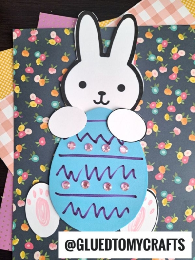 A cute bunny craft made of paper, holding a decorated blue foam egg craft with sequins and squiggly lines, placed on floral and plaid patterned paper. @GLUEDTOMYCRAFTS is written at the bottom.