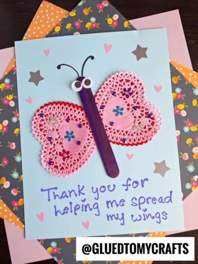 A handmade card features a butterfly crafted from a purple popsicle stick, pink paper doilies, googly eyes, and heart stickers on blue paper. Perfect for fans of Paper Doily Crafts, it reads, “Thank you for helping me spread my wings.”.