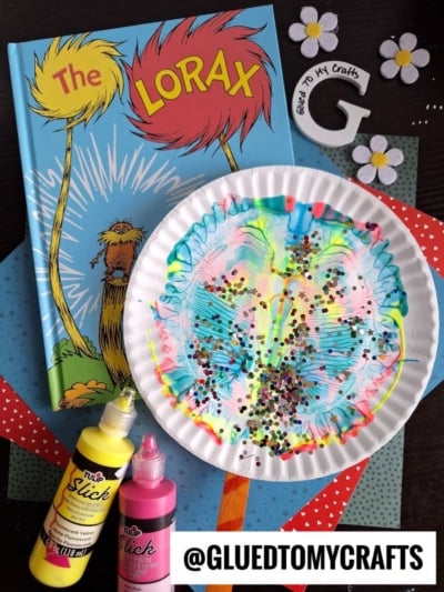 A colorful craft scene with āThe Loraxā book, a painted paper plate art featuring paint splat truffula trees, sparkling sequins, bottles of yellow and pink paint, polka dot paper, foam flowers, and a wooden letter āG.ā Text: @GLUEDTOMYCRAFTS.