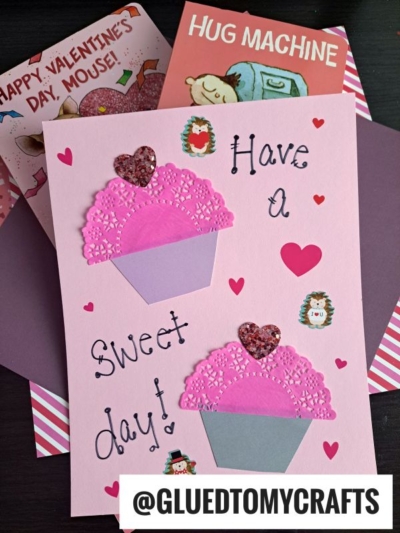 A handmade Valentine’s card featuring paper doily cupcake cards on pink paper, decorated with glitter hearts and pink doilies, placed atop two illustrated Valentine’s cards. The handle @GLUEDTOMYCRAFTS appears at the bottom.