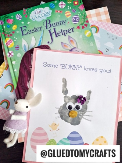 A children’s book titled Easter Bunny Helper, pastel cardstock, a bunny plush, and a handprint bunny craft card with googly eyes, pom-pom nose, and Some BUNNY loves you! text are arranged together.