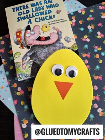 A children’s book titled There Was an Old Lady Who Swallowed a Chick! lies on floral paper, next to a yellow foam egg craft with googly eyes and an orange beak. The handle @GLUEDTOMYCRAFTS appears at the bottom.