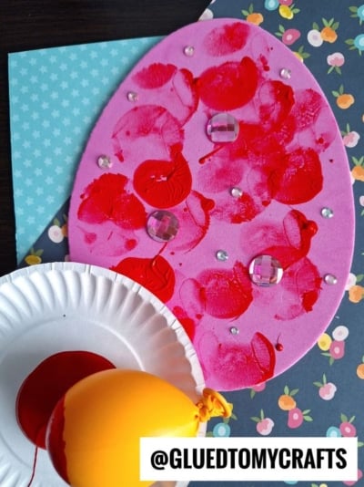 A pink foam egg craft decorated with red paint prints, clear gems, and rhinestones sits on patterned paper. A yellow balloon and a paper plate with red paint are nearby. Text reads @GLUEDTOMYCRAFTS.