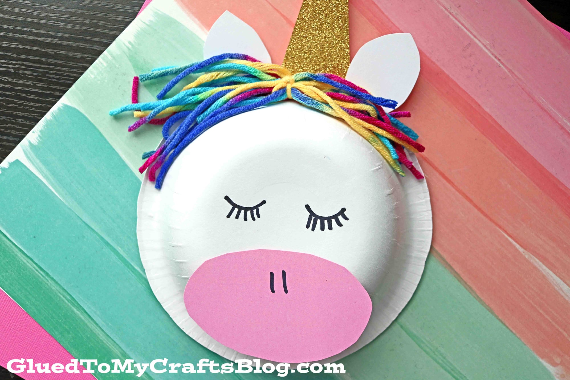 A vibrant paper bowl unicorn features a gold glitter horn, white ears, rainbow yarn mane, drawn eyes and nostrils, and a pink paper snout on a striped background.